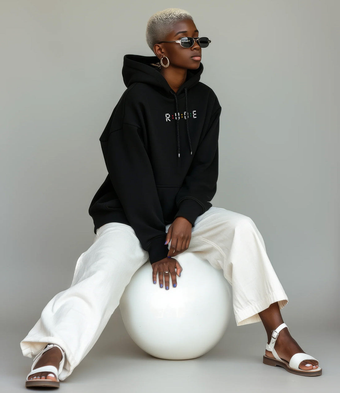 Woman wearing Rude Attire black hoodie with R·U·D·E logo, seated on a white sphere, styled with wide-leg cream trousers and white sandals